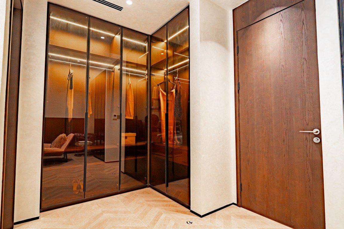 Walk-in closet with glass-panel doors and clothes hanging on racks, adjacent to a tall wooden door, warm lighting