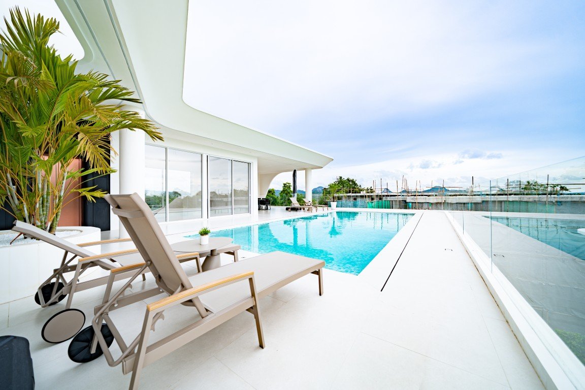 Modern rooftop pool area with white loungers, glass railing, and a palm plant in view.