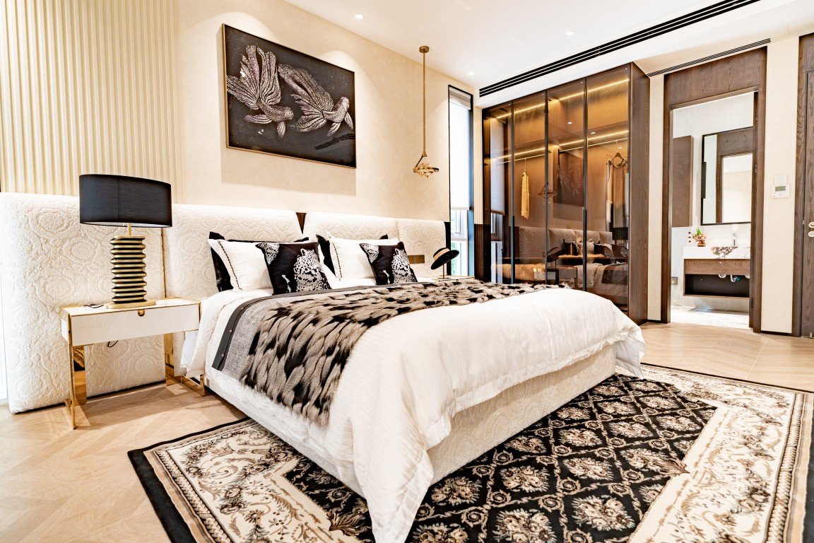 Luxurious modern bedroom featuring a king-size bed with an ornate tufted headboard, white bedding, and a black-and-white fur throw, plus gold-accented nightstands and a large decorative rug.