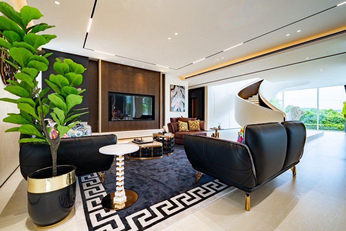 Luxurious modern living room with black leather sofas, a dark wood accent wall with a TV, and a white spiral staircase by large windows.