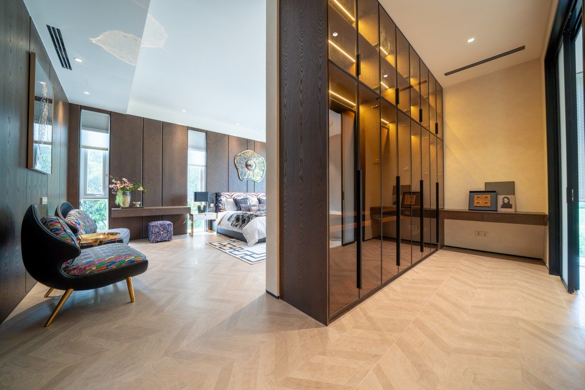 Open-plan modern suite: a seating area with colorful chairs, a bed in the back, and a large glass-front wardrobe along a dark wood wall on the right.