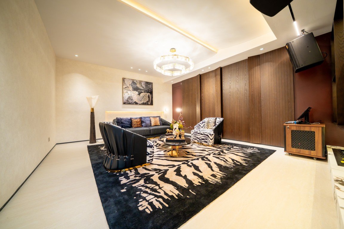 Modern living room with zebra-pattern rug, black seating, and warm lighting; wooden paneled wall and a circular chandelier center-stage.
