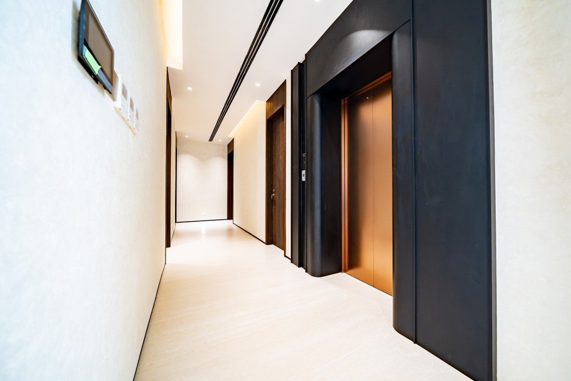 Long modern hotel hallway with multiple doors and an open elevator bank on the right, warm copper elevator doors, and bright recessed lighting.