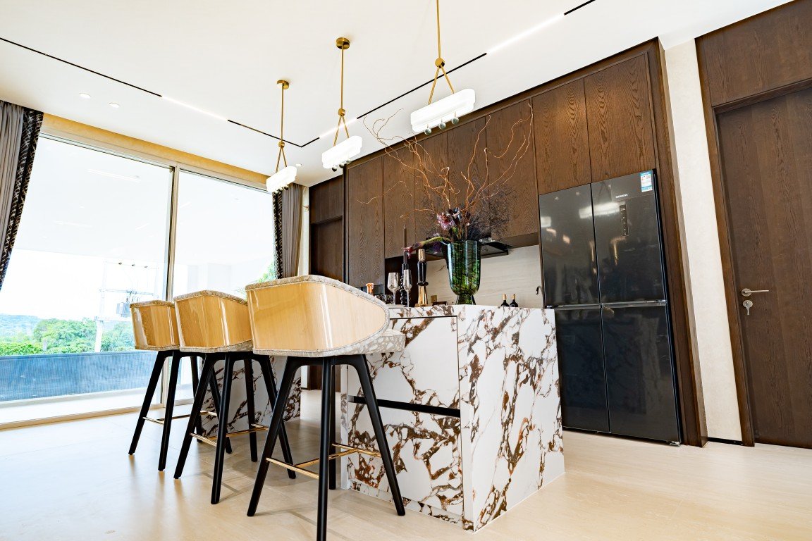 Contemporary kitchen with a marble island, four beige bar stools, and dark wood cabinets beneath pendant lights.