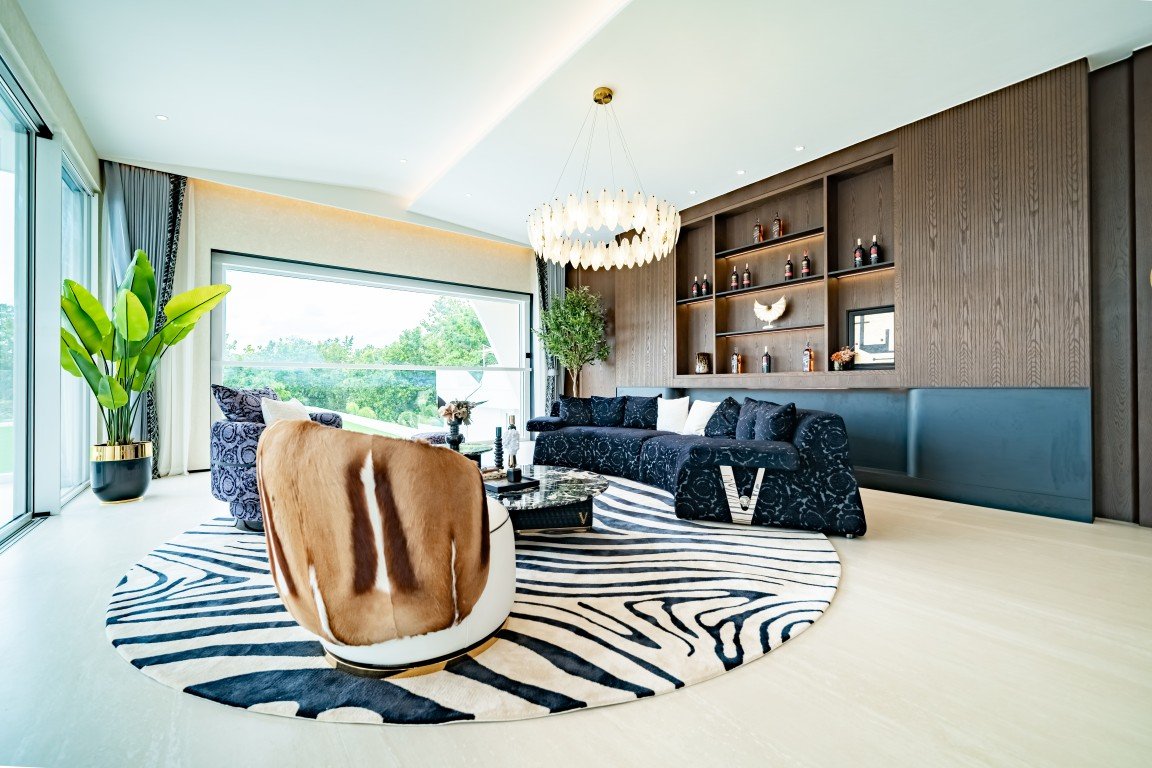 Modern living room with a zebra-pattern rug, dark sofa, and a curved brown chair under a sculptural chandelier. A large window overlooks greenery and a plant in the corner adds life to the space, along with built-in shelving on the wall.