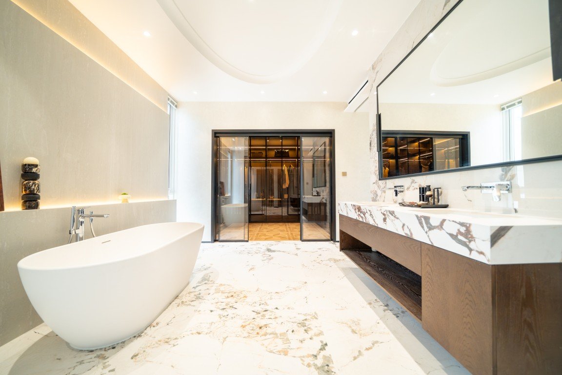 Luxurious spa-like bathroom with a freestanding oval tub, marble countertops, and double sinks beside a large mirror. A glass-door closet is in the background.