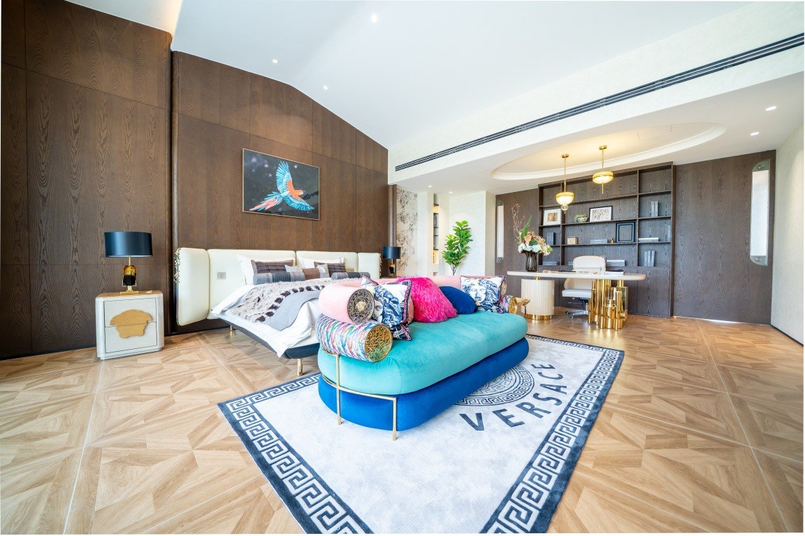 Luxurious hotel suite living area with a white bed, teal sofa, and a Versace rug on a herringbone wood floor.