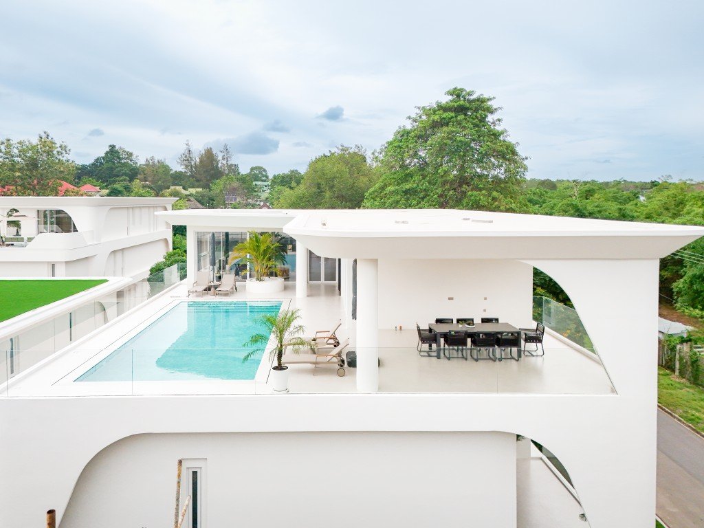 Modern white rooftop terrace with rectangular pool, lounge chairs, and glass railing overlooking green trees.