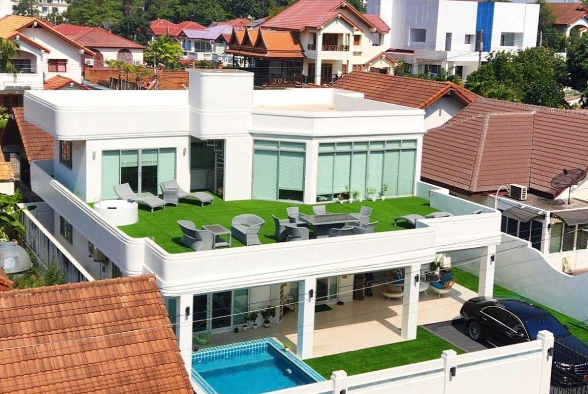 Modern white villa with a rooftop lawn and outdoor seating area overlooking neighboring houses and red-tiled roofs; in-ground pool visible on lower level.
