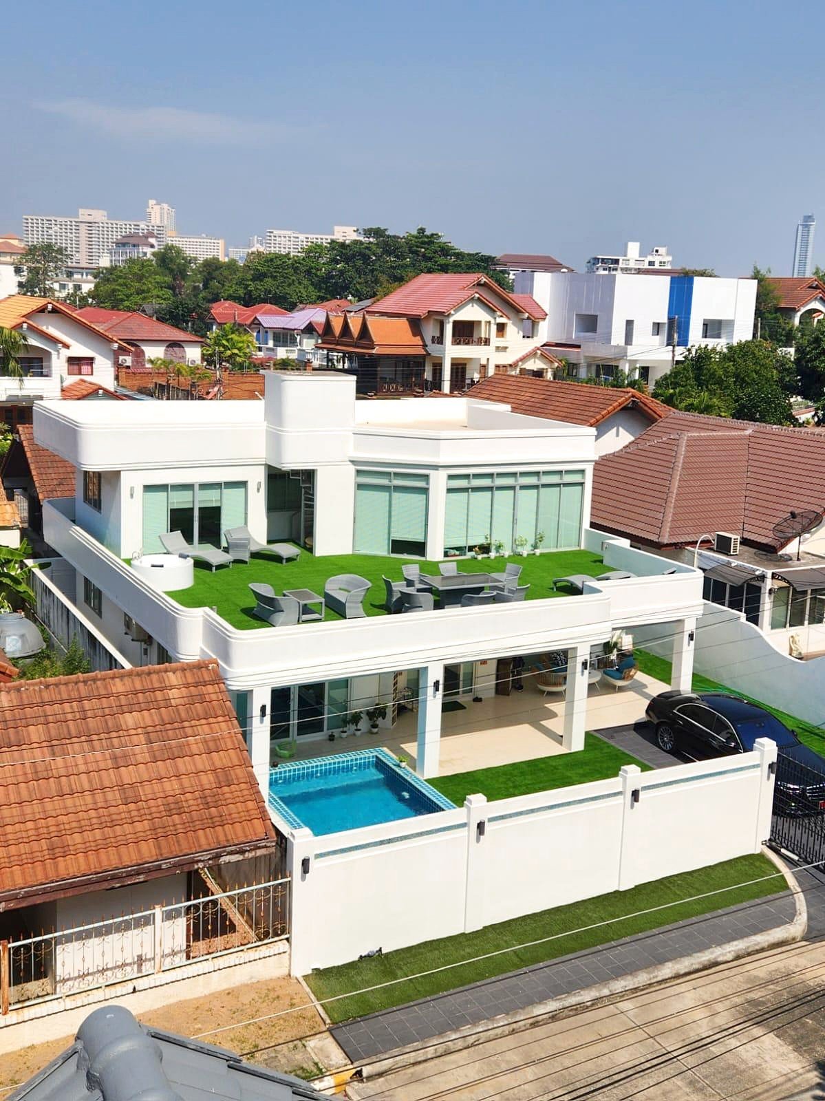 Casa_Jomtien_Village_Rent_Rdl346 Modern white villa with a rooftop lawn and outdoor seating area overlooking neighboring houses and red-tiled roofs; in-ground pool visible on lower level.