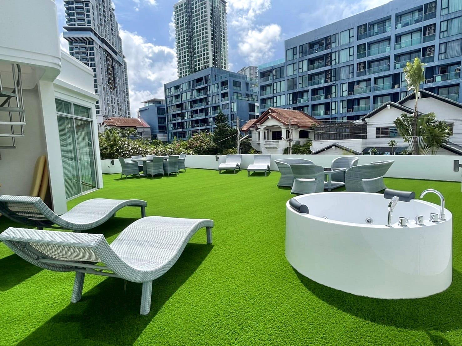 Casa_Jomtien_Village_Rent_Rdl346_10 Rooftop terrace with bright green artificial grass, white lounge chairs, and a circular white hot tub, city buildings in the background