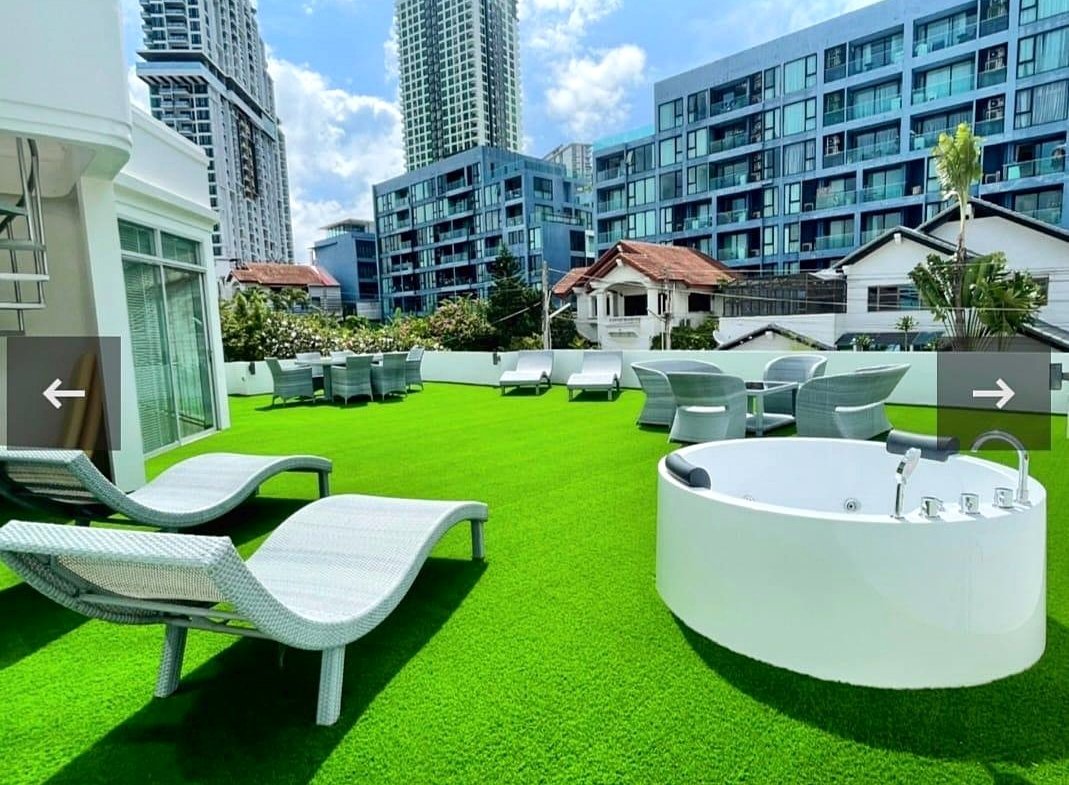 Rooftop terrace with white lounge chairs, a round white hot tub, and outdoor dining tables on bright green artificial grass, urban high-rise buildings in the background.