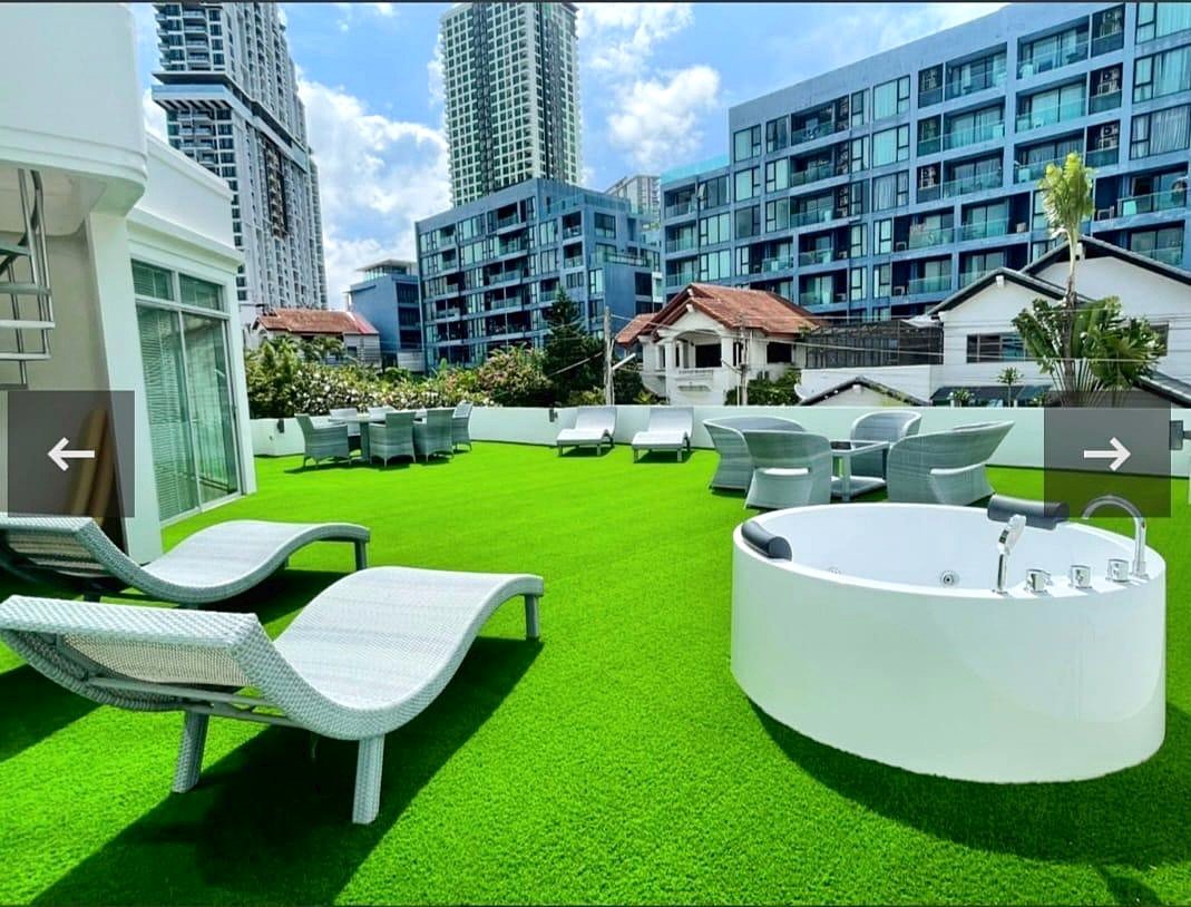 Casa_Jomtien_Village_Rent_Rdl346_11 Rooftop terrace with white lounge chairs, a round white hot tub, and outdoor dining tables on bright green artificial grass, urban high-rise buildings in the background.