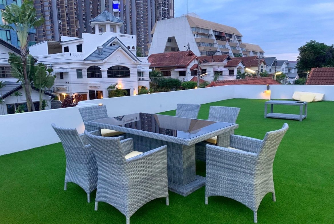 Rooftop terrace with gray wicker dining set on artificial green grass, white walls, and city buildings in the background.