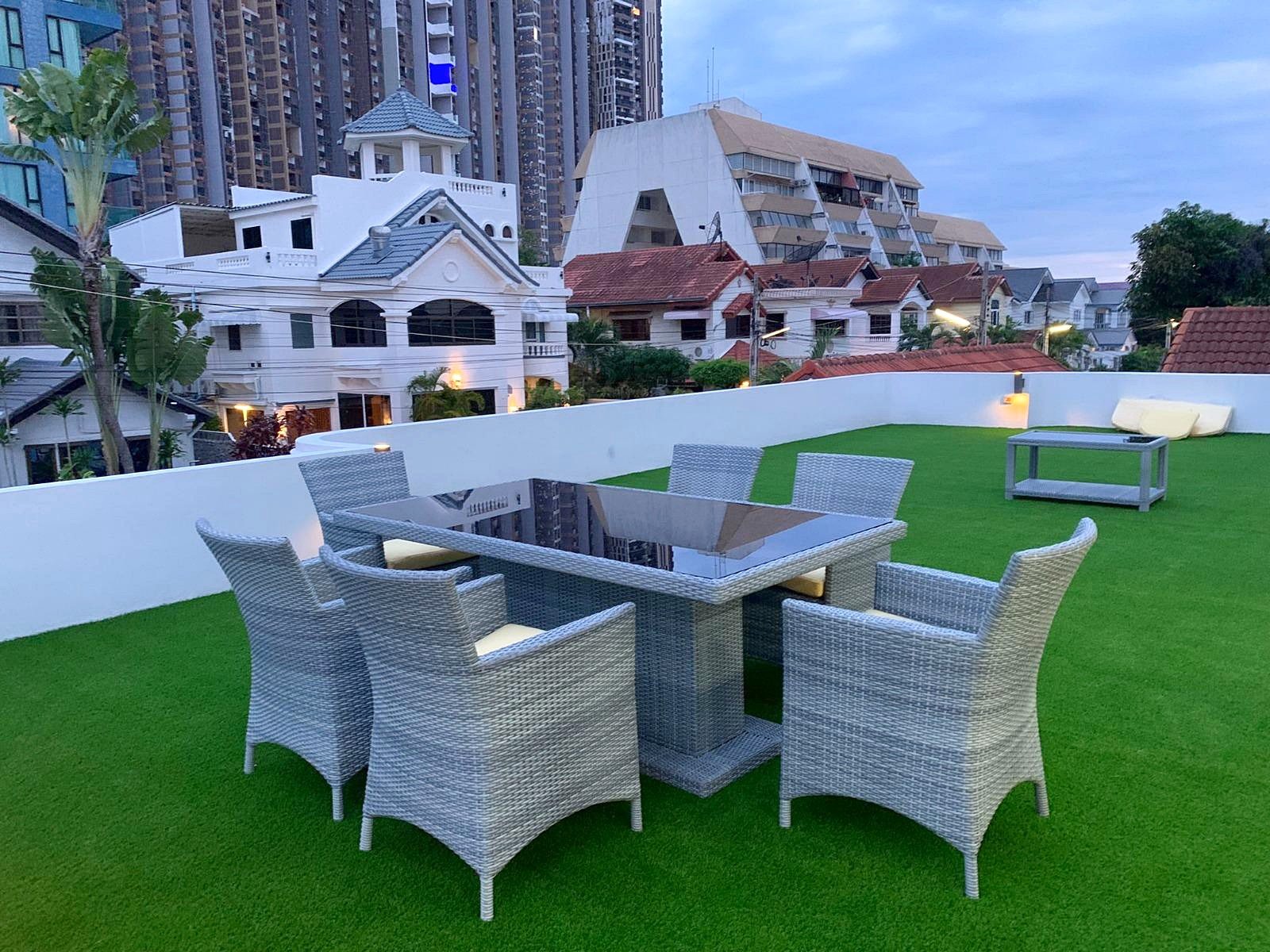 Casa_Jomtien_Village_Rent_Rdl346_13 Rooftop terrace with gray wicker dining set on artificial green grass, white walls, and city buildings in the background.