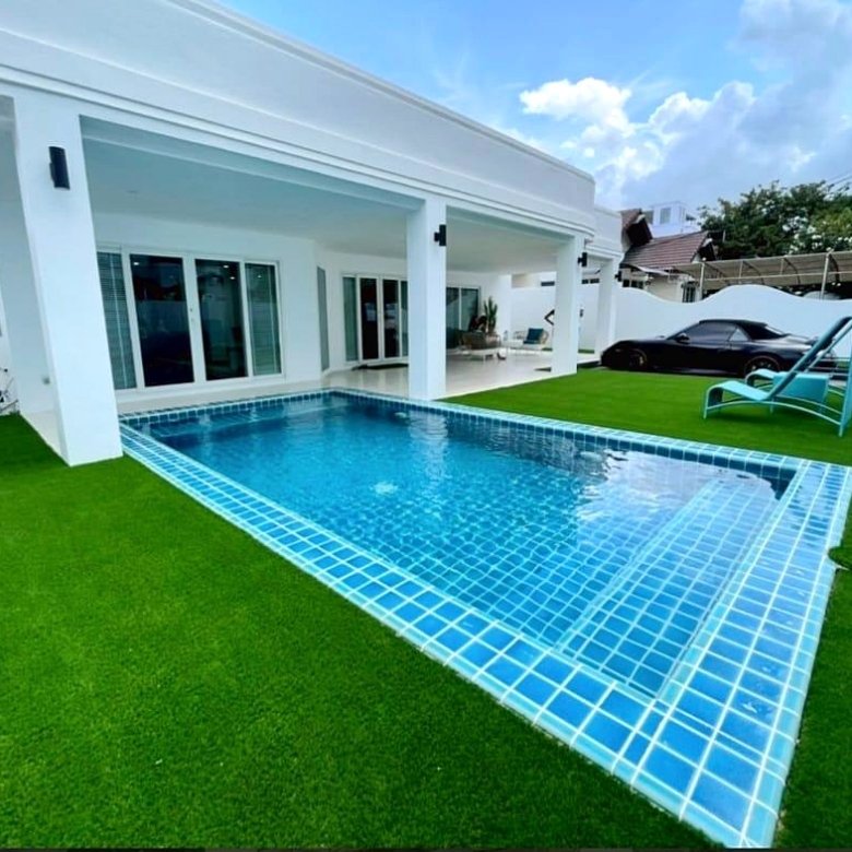 Backyard with a rectangular blue tiled pool white house and green artificial grass a black car sits on the driveway in the background