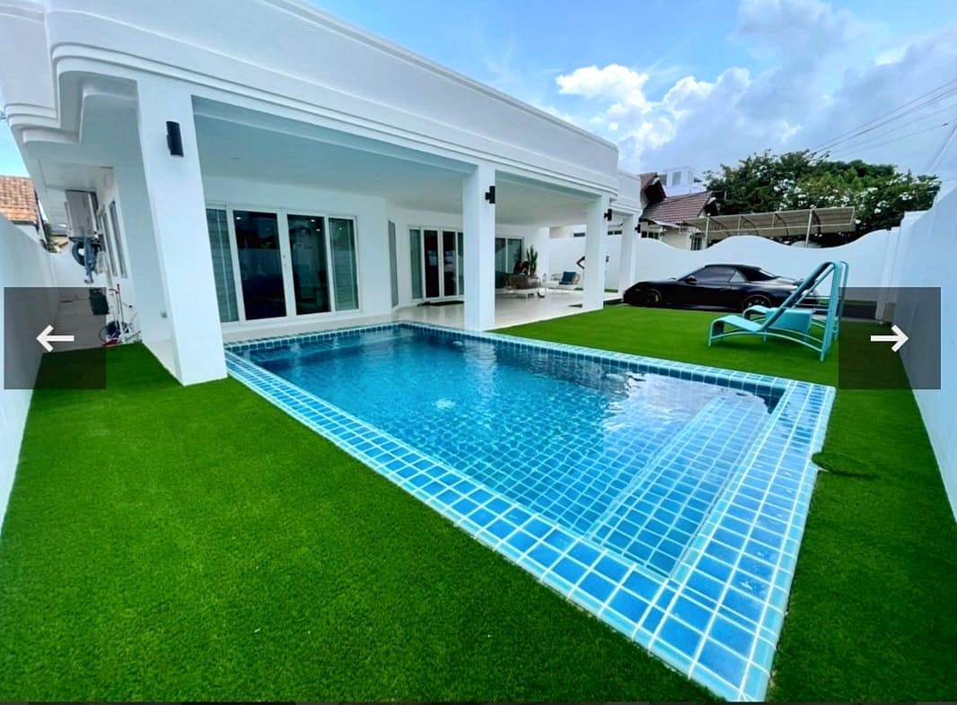 Backyard with a rectangular blue tiled pool white house and green artificial grass a black car sits on the driveway in the background