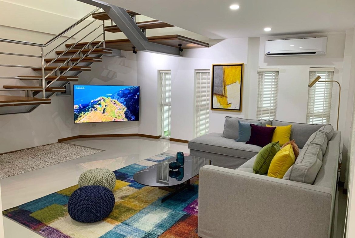 Modern living room with a gray sectional, colorful cushions, glass coffee table, and a wall-mounted TV under a staircase.