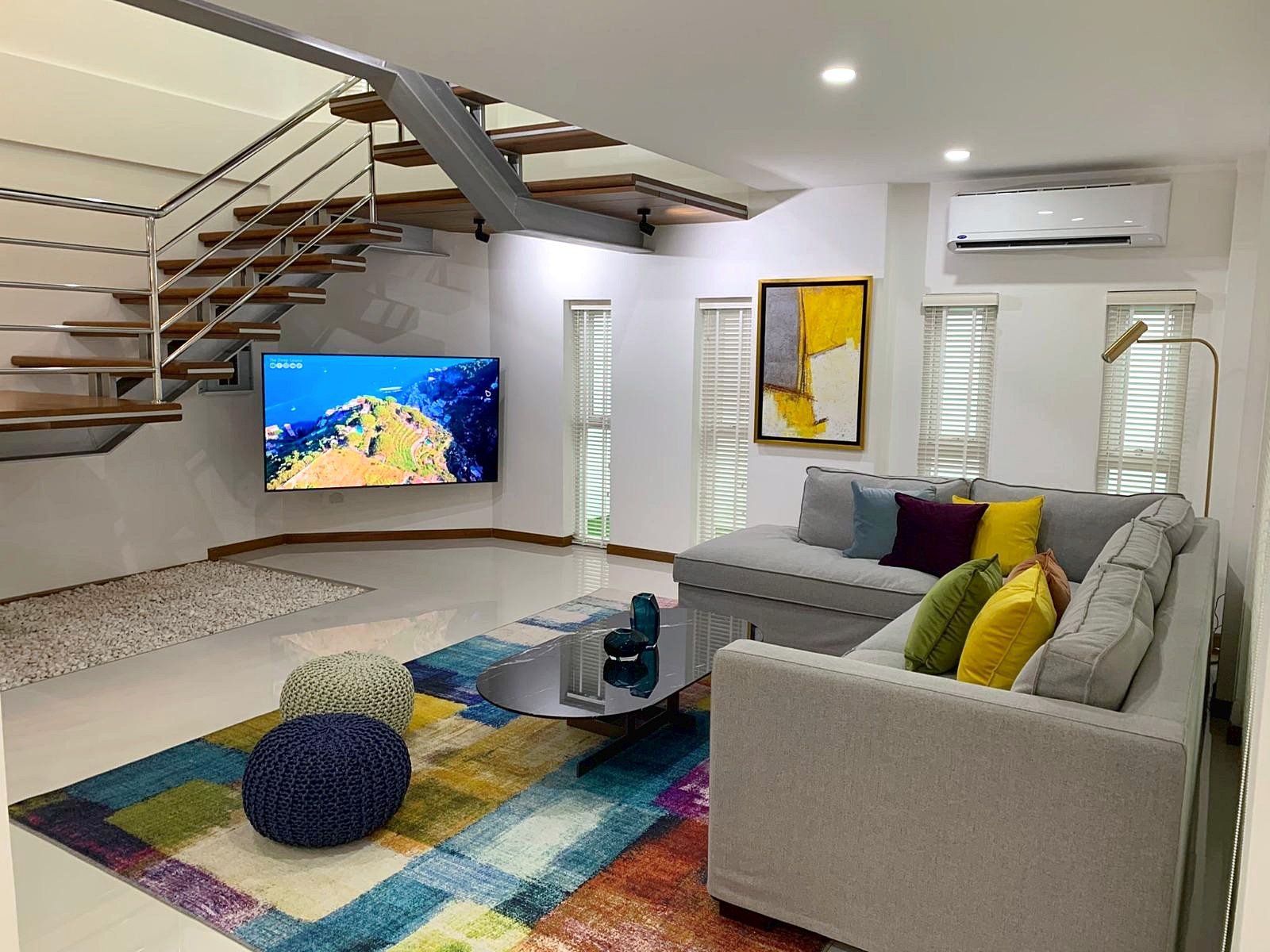 Casa_Jomtien_Village_Rent_Rdl346_3 Modern living room with a gray sectional, colorful cushions, glass coffee table, and a wall-mounted TV under a staircase.