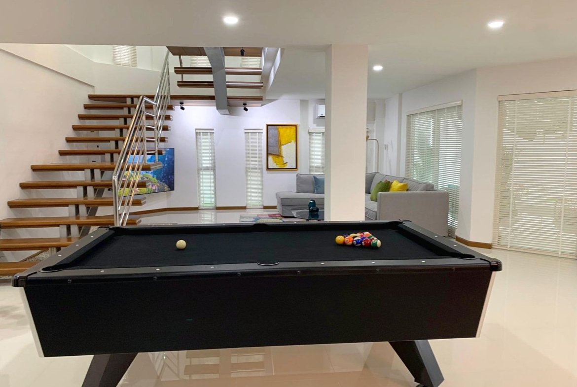 Modern basement recreation room with a black pool table in foreground, chalk balls racked on the right, and a wooden staircase on the left.