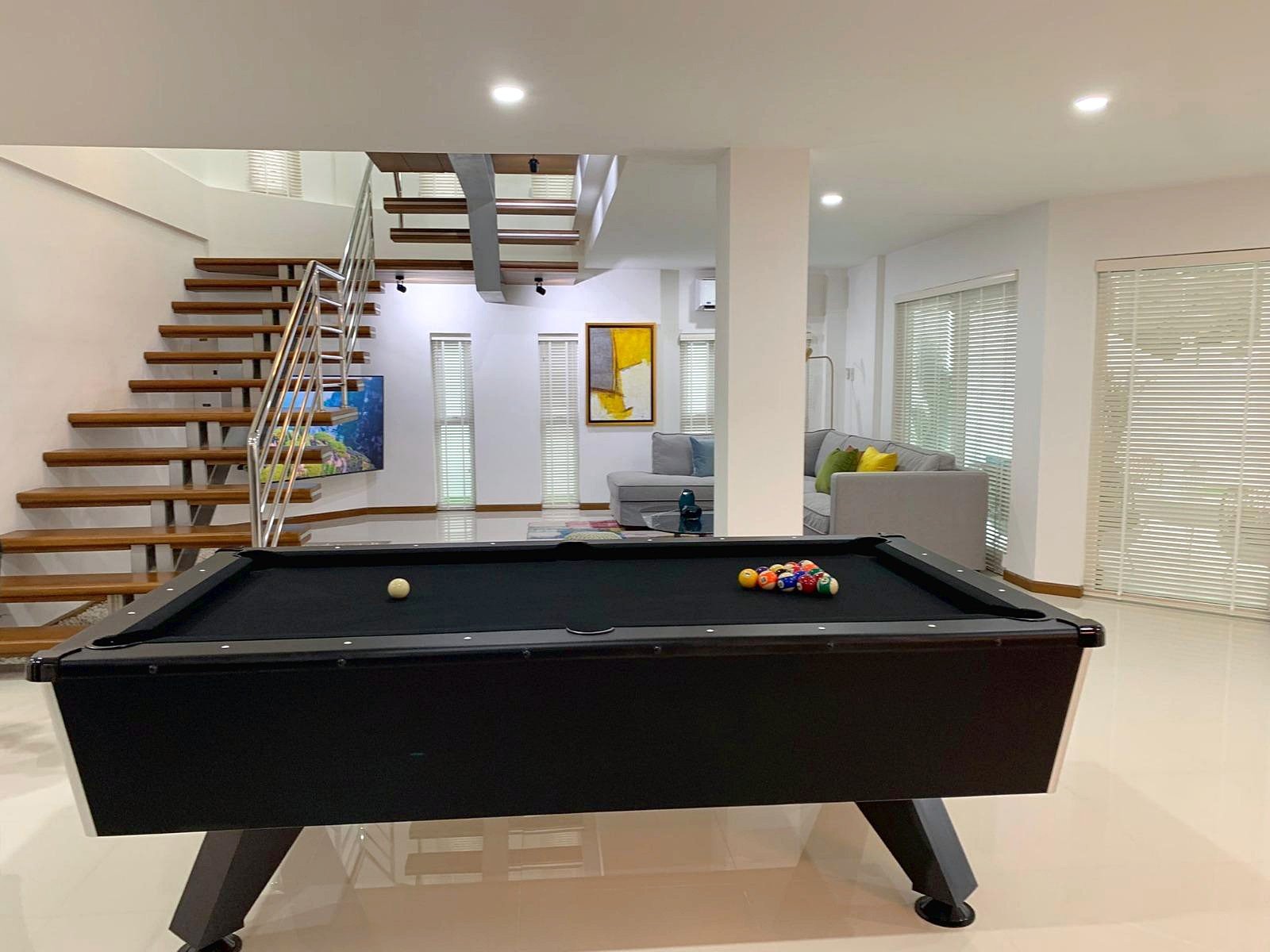 Casa_Jomtien_Village_Rent_Rdl346_4 Modern basement recreation room with a black pool table in foreground, chalk balls racked on the right, and a wooden staircase on the left.