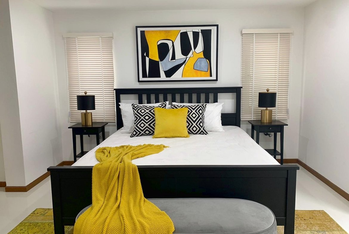 Dark wooden bed with white bedding, yellow throw, and black‑and‑white geometric pillows in a modern bedroom, abstract yellow/black art above the headboard.