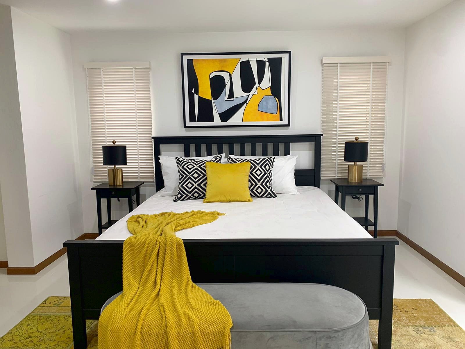 Casa_Jomtien_Village_Rent_Rdl346_5 Dark wooden bed with white bedding, yellow throw, and black‑and‑white geometric pillows in a modern bedroom, abstract yellow/black art above the headboard.