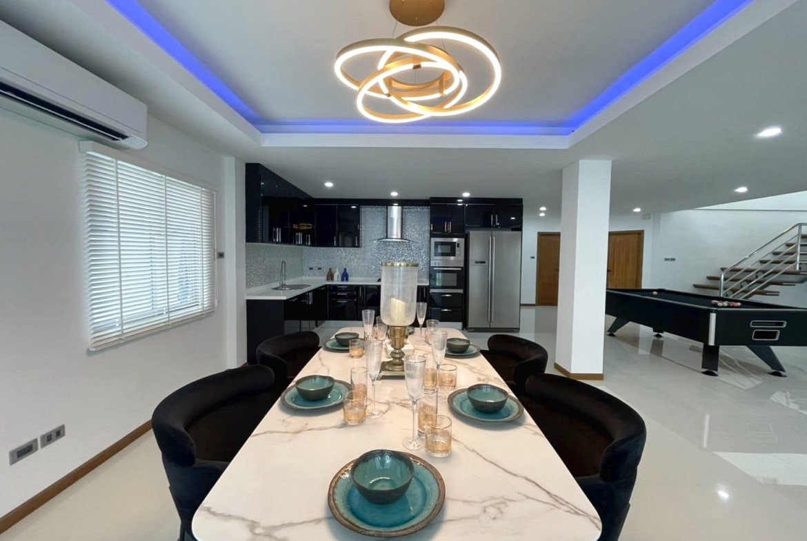 Open-plan kitchen and dining area with a marble dining table set for eight, teal bowls and glassware, and a modern circular chandelier under blue ceiling lighting.