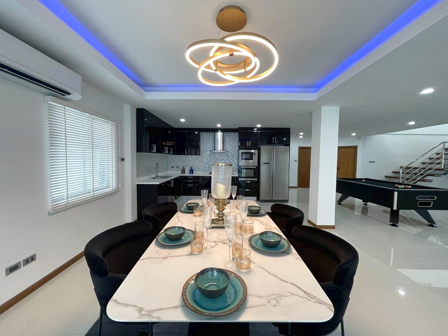 Casa_Jomtien_Village_Rent_Rdl346_6 Open-plan kitchen and dining area with a marble dining table set for eight, teal bowls and glassware, and a modern circular chandelier under blue ceiling lighting.