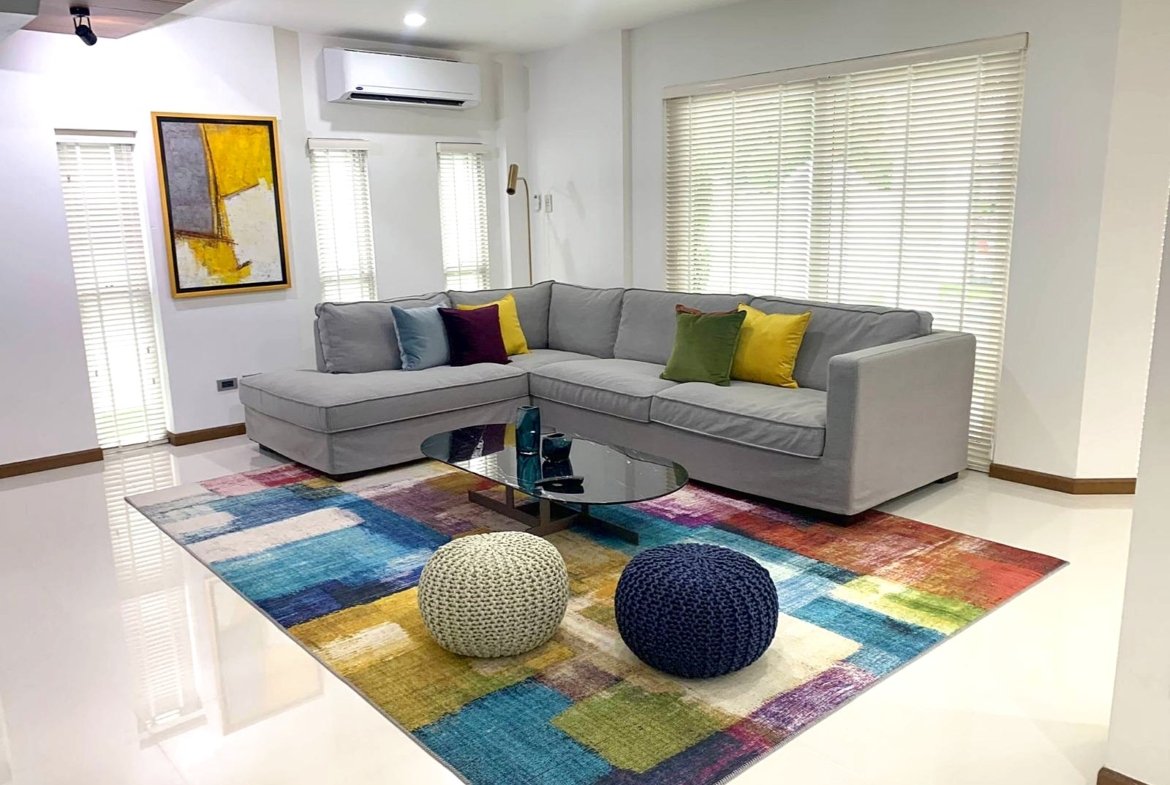 Bright living room with a light gray L-shaped sofa, colorful cushions, a glass coffee table, and a multicolored rug under two knitted poufs.