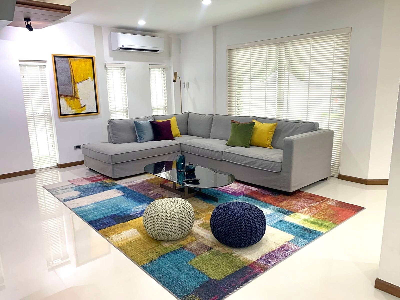 Casa_Jomtien_Village_Rent_Rdl346_7 Bright living room with a light gray L-shaped sofa, colorful cushions, a glass coffee table, and a multicolored rug under two knitted poufs.