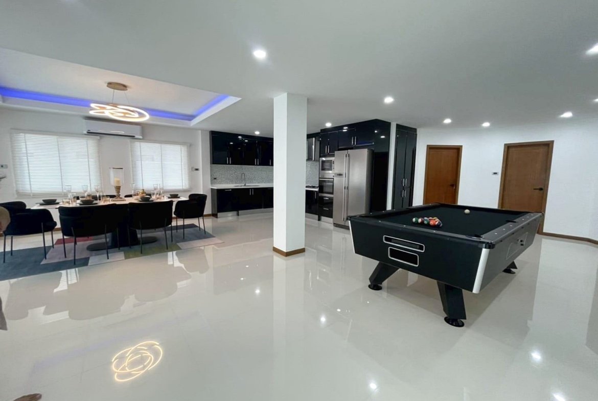 Open-concept living space with a black pool table in the foreground, a white glossy floor, and a modern kitchen with stainless steel appliances in the background.