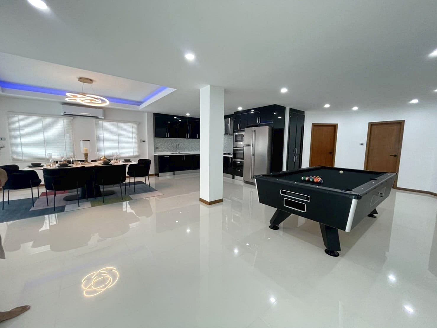 Casa_Jomtien_Village_Rent_Rdl346_8 Open-concept living space with a black pool table in the foreground, a white glossy floor, and a modern kitchen with stainless steel appliances in the background.