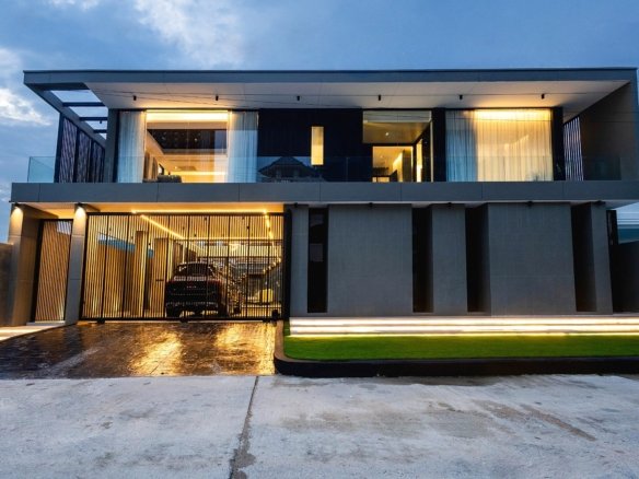 Modern twostory house with glass balcony grey facade and warm interior lighting visible through large windows a gated driveway with a parked car under ambient exterior lights at dusk