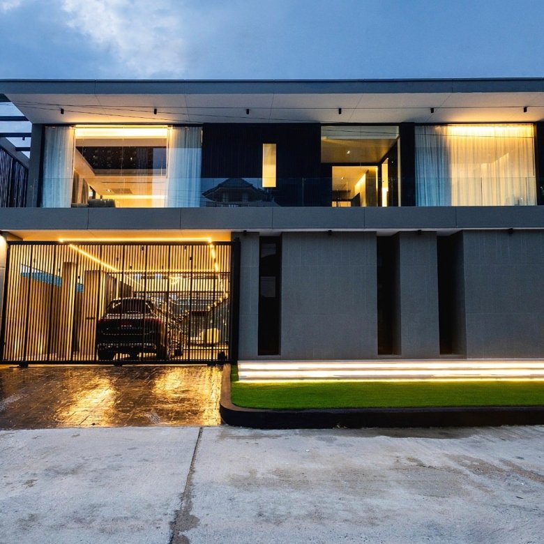 Modern twostory house with glass balcony grey facade and warm interior lighting visible through large windows a gated driveway with a parked car under ambient exterior lights at dusk
