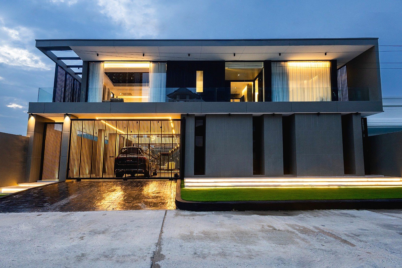 Casa_Jomtien_Village_Rent_Rdl352 Modern two‑story house with glass balcony, grey facade, and warm interior lighting visible through large windows; a gated driveway with a parked car under ambient exterior lights at dusk.