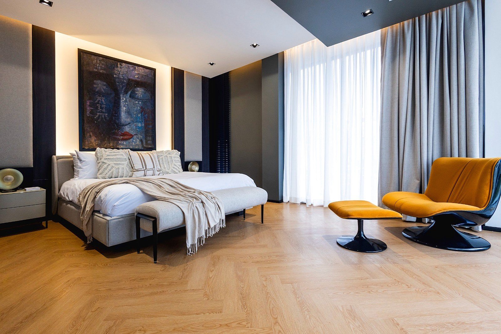 Casa_Jomtien_Village_Rent_Rdl352_11 Modern bedroom with a beige upholstered bed, neutral bedding, and a matching bench at the foot of the bed, warm wood floor, and an abstract blue portrait above the headboard.