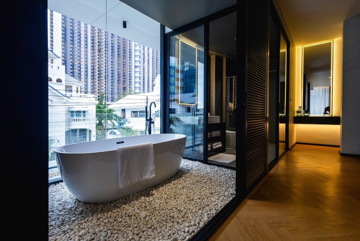 Spacious bathroom with a freestanding white bathtub on a pebble floor by a large window overlooking city skyscrapers