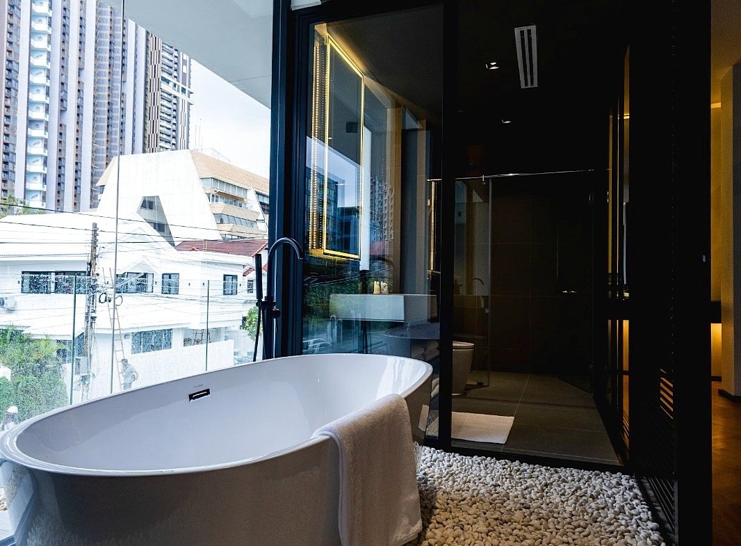 Freestanding white bathtub on a pebble floor beside a large glass wall with city buildings outside, modern bathroom setting