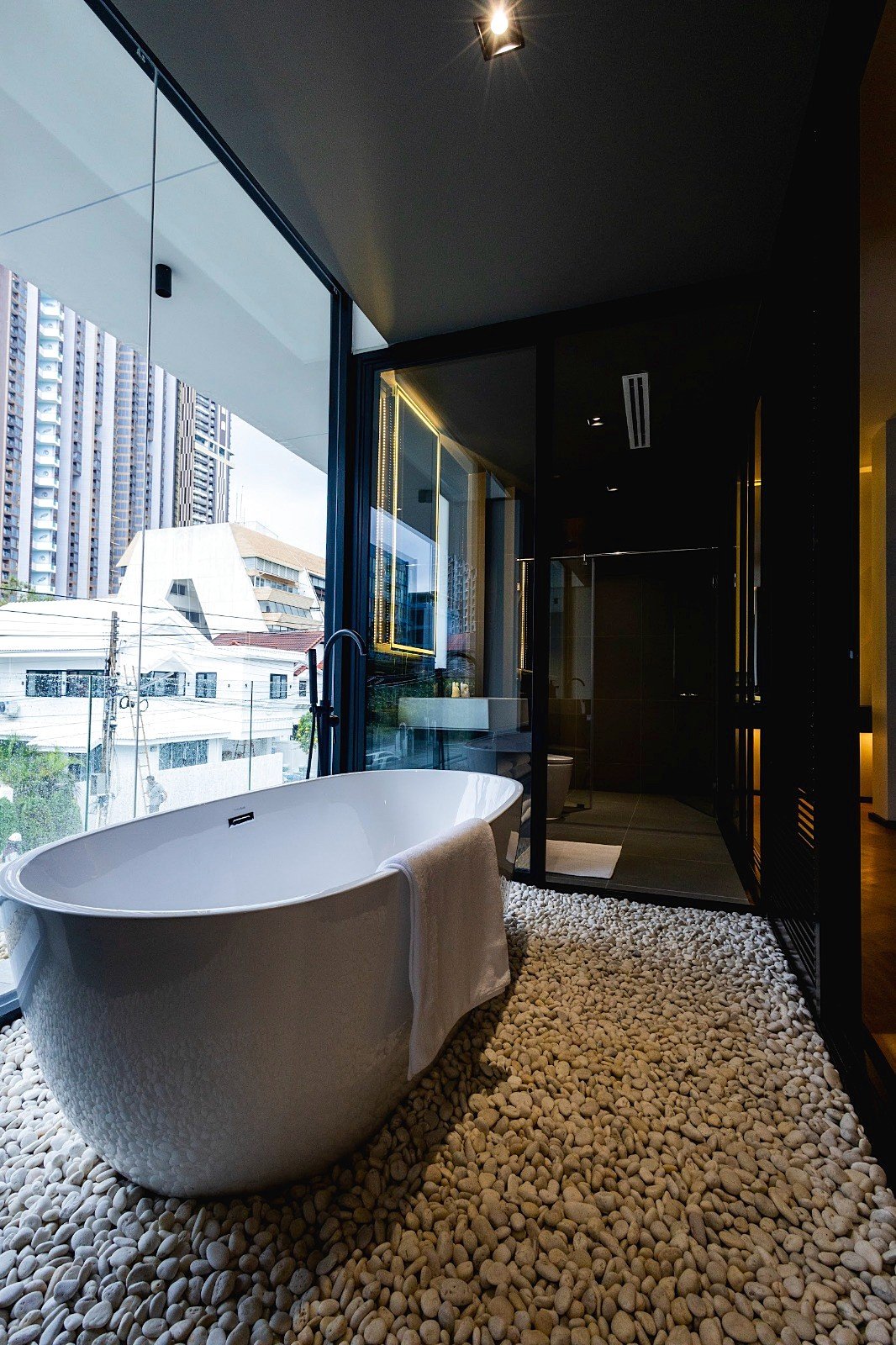 Casa_Jomtien_Village_Rent_Rdl352_18 Freestanding white bathtub on a pebble floor beside a large glass wall with city buildings outside, modern bathroom setting