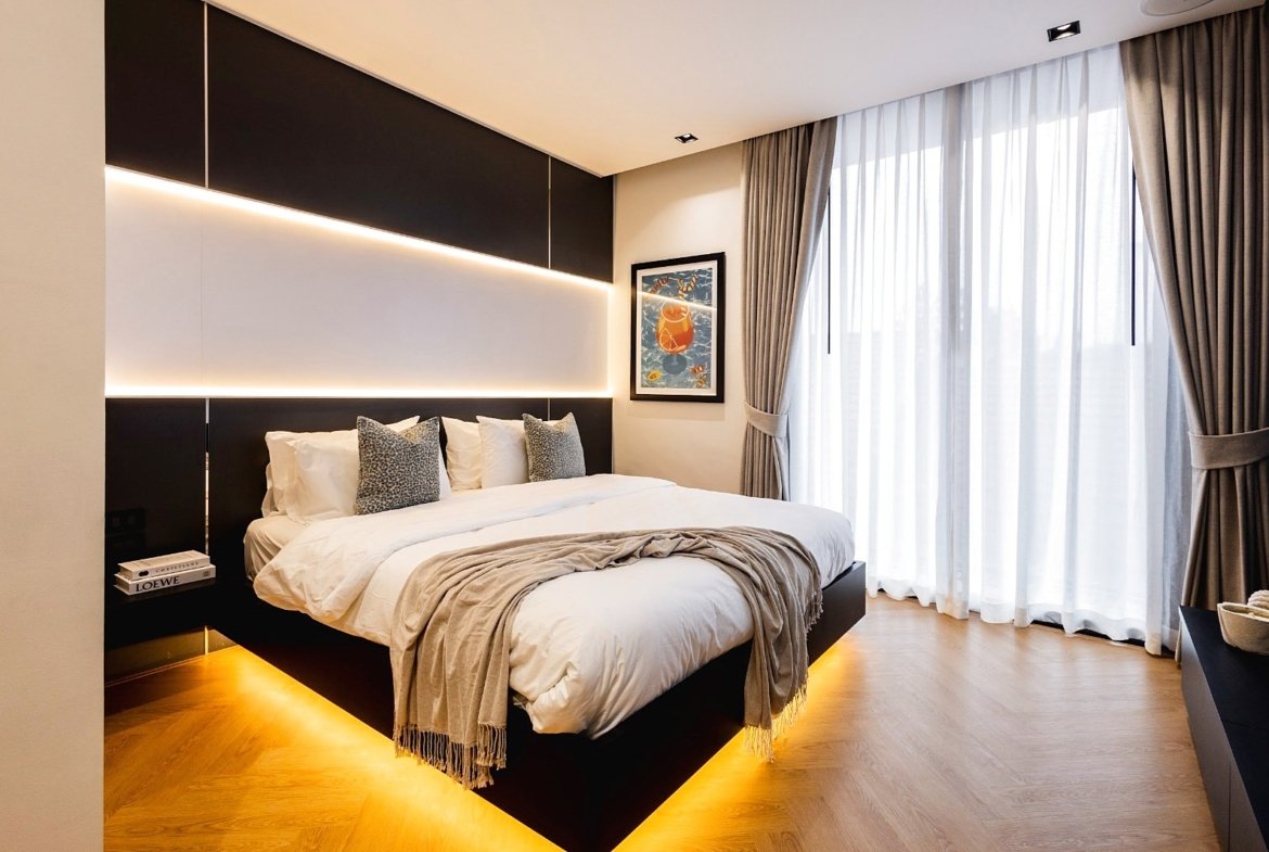 Modern hotel bedroom with a low black platform bed, white bedding, grey throw, and backlit headboard panels head of a warm glow from the lighting system.