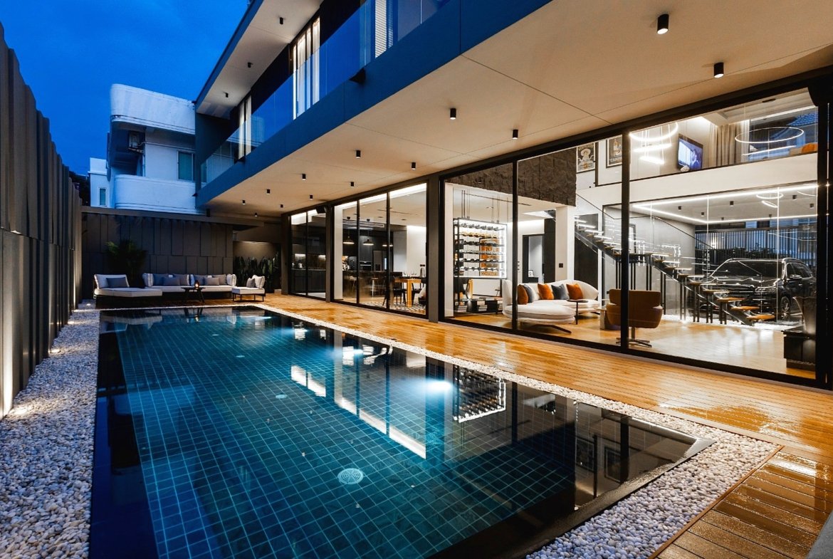 Modern residence at dusk featuring a rectangular pool, wooden deck, and floor-to-ceiling glass walls revealing a stylish interior.