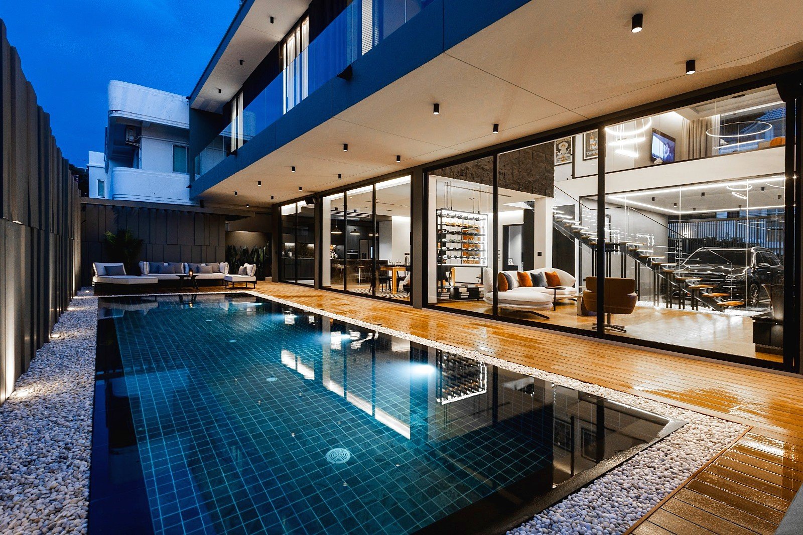 Casa_Jomtien_Village_Rent_Rdl352_2 Modern residence at dusk featuring a rectangular pool, wooden deck, and floor-to-ceiling glass walls revealing a stylish interior.