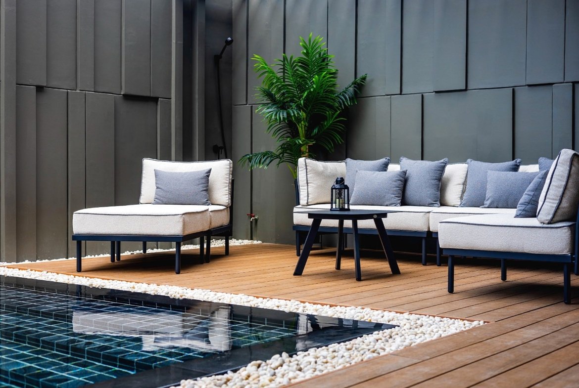 Beige outdoor sectional with blue-gray cushions on a wooden deck, next to a round black table and a tall green plant against a dark wall.