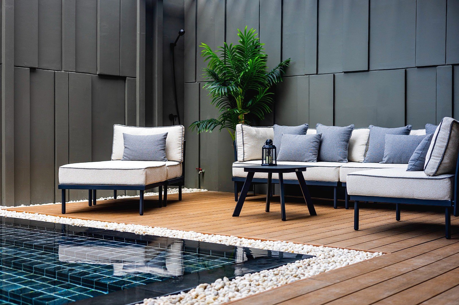Casa_Jomtien_Village_Rent_Rdl352_28 Beige outdoor sectional with blue-gray cushions on a wooden deck, next to a round black table and a tall green plant against a dark wall.
