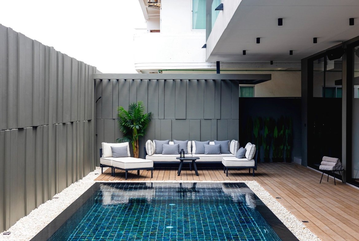 Modern outdoor lounge area with a gray sectional sofa on a wooden deck beside a rectangular pool and a tall gray privacy wall.
