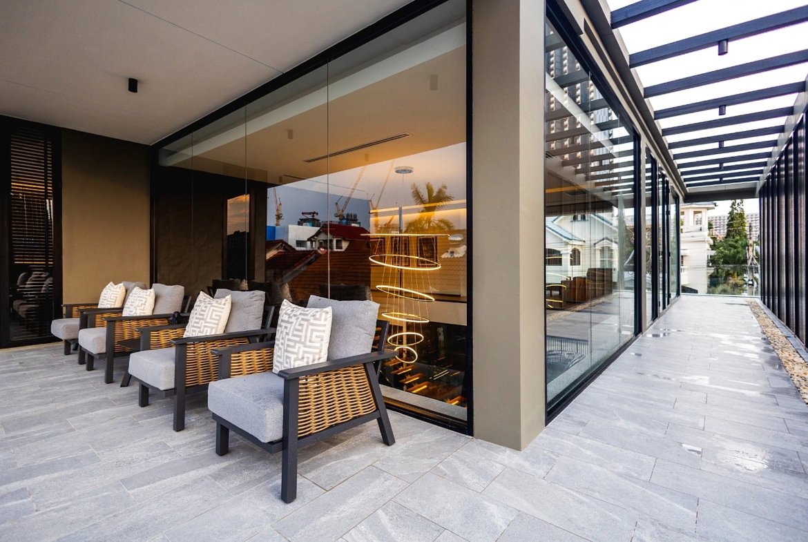 Modern outdoor lounge with wicker chairs and grey cushions along a glass-walled building exterior at dusk. Reflective glass shows a spiral pendant light inside and city beyond.