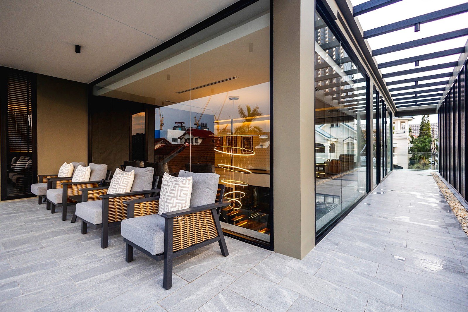 Casa_Jomtien_Village_Rent_Rdl352_30 Modern outdoor lounge with wicker chairs and grey cushions along a glass-walled building exterior at dusk. Reflective glass shows a spiral pendant light inside and city beyond.