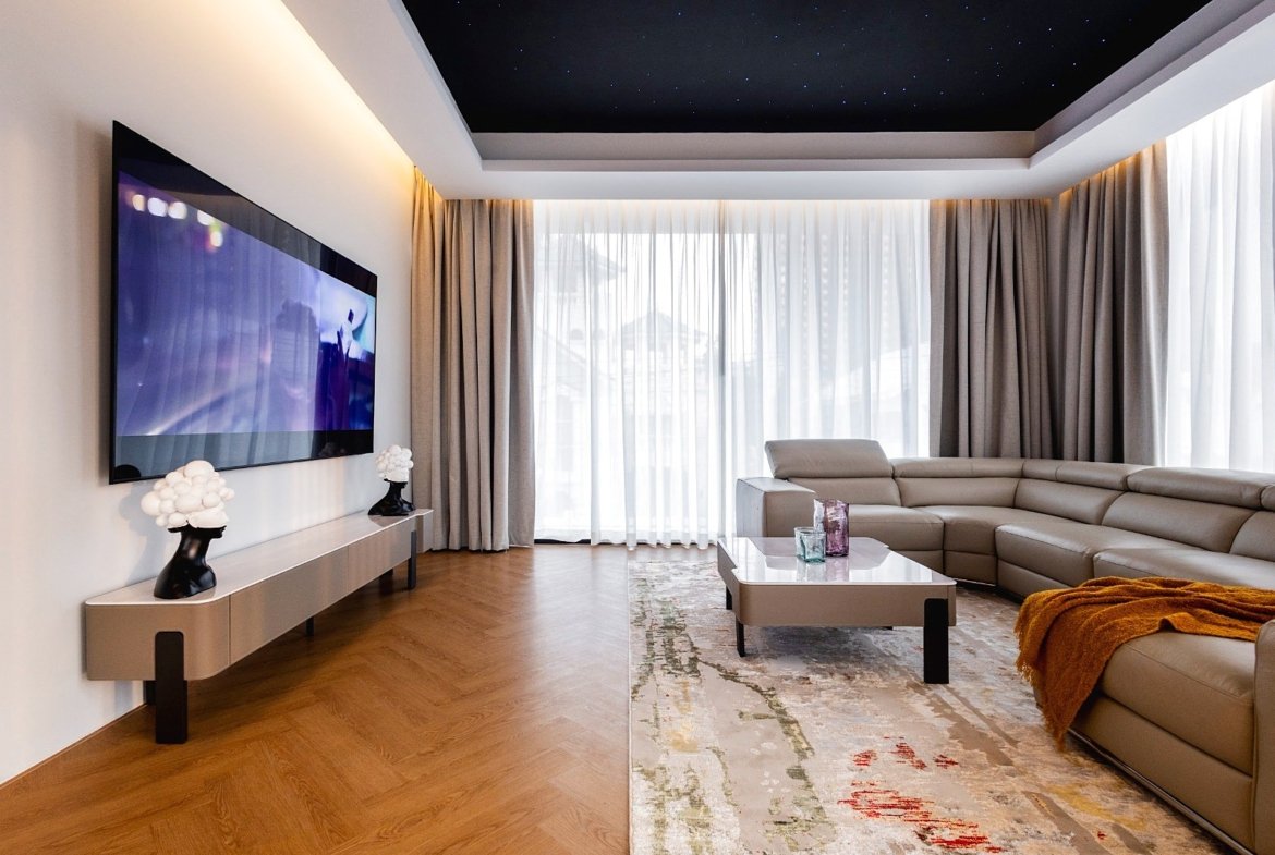 Modern living room with a wall-mounted TV, beige sectional sofa, and glass-top coffee table under a starry ceiling.