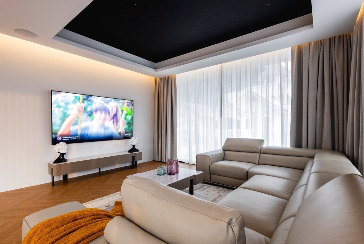 A modern living room with an L-shaped beige leather sofa facing a wall-mounted flat-screen TV and a low console table with decorative lamps on either side.